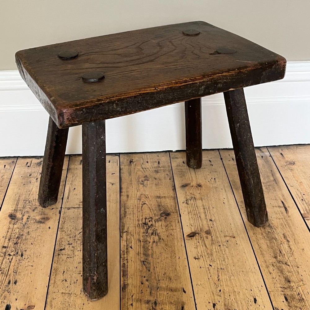A rustic dark oak four-legged stool featuring a thick rectangular top with four visible circular wedged tenons and splayed faceted legs.