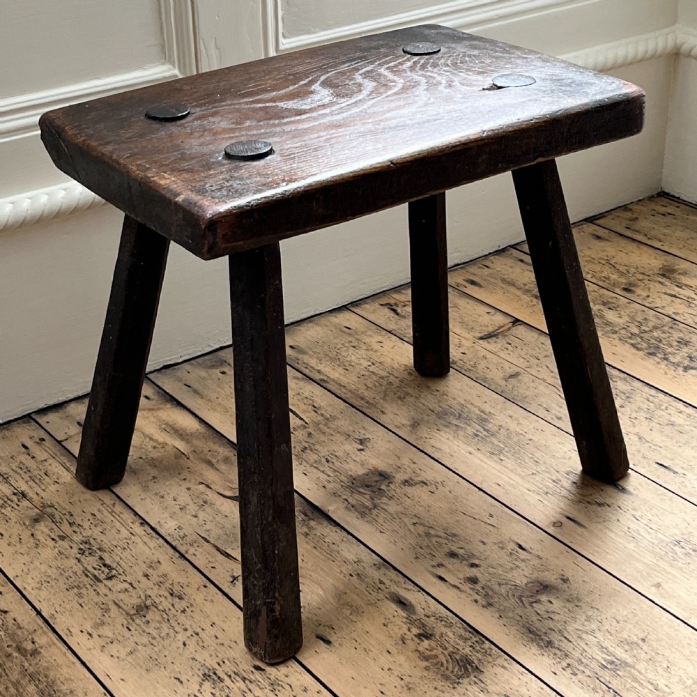 A rustic dark oak four-legged stool featuring a thick rectangular top with four visible circular wedged tenons and splayed faceted legs. View II