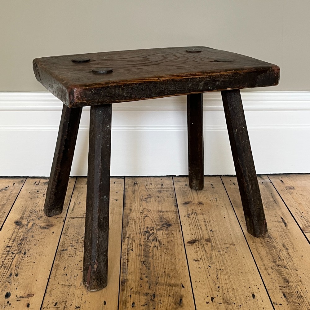 A rustic dark oak four-legged stool featuring a thick rectangular top with four visible circular wedged tenons and splayed faceted legs. View III