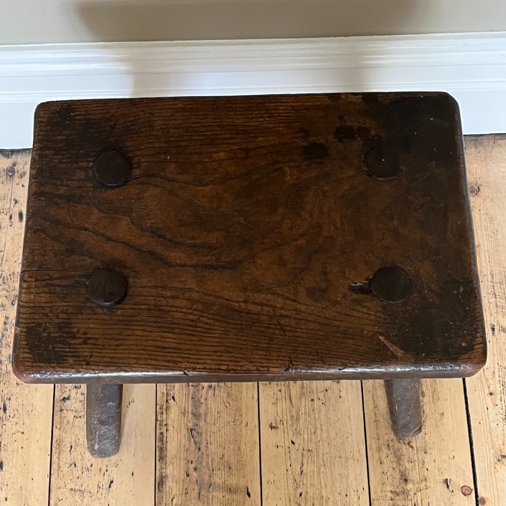 A rustic dark oak four-legged stool featuring a thick rectangular top with four visible circular wedged tenons and splayed faceted legs. View V