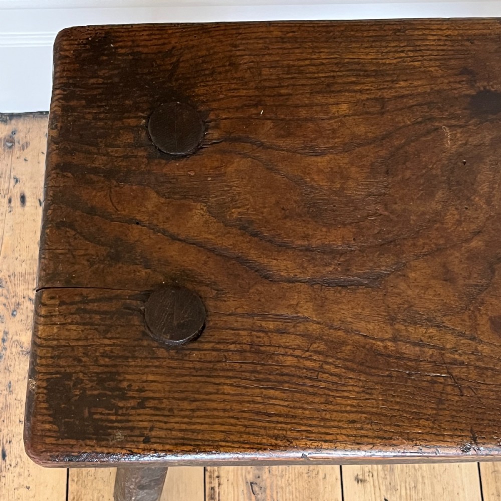 A rustic dark oak four-legged stool featuring a thick rectangular top with four visible circular wedged tenons and splayed faceted legs. View VI