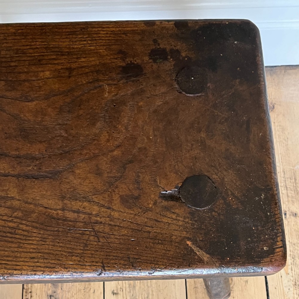 A rustic dark oak four-legged stool featuring a thick rectangular top with four visible circular wedged tenons and splayed faceted legs. View VII