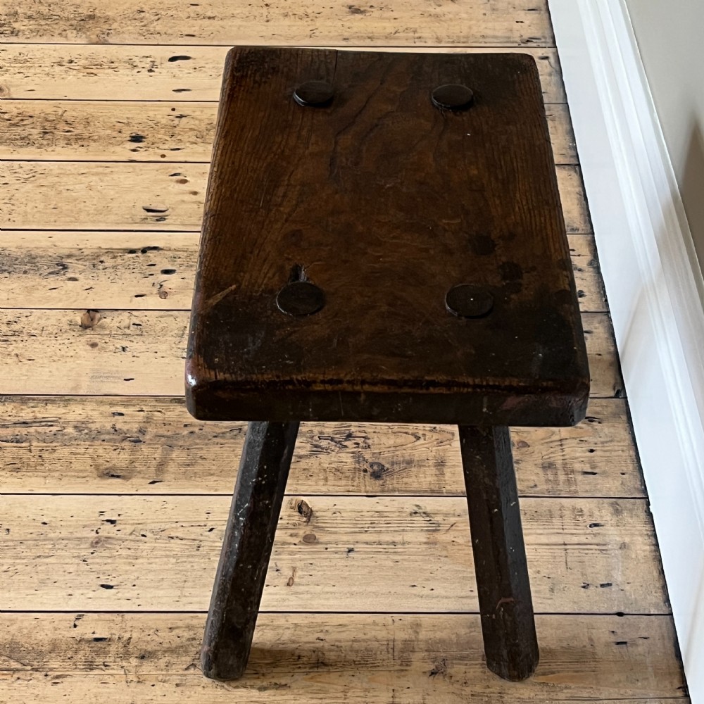 A rustic dark oak four-legged stool featuring a thick rectangular top with four visible circular wedged tenons and splayed faceted legs. View X
