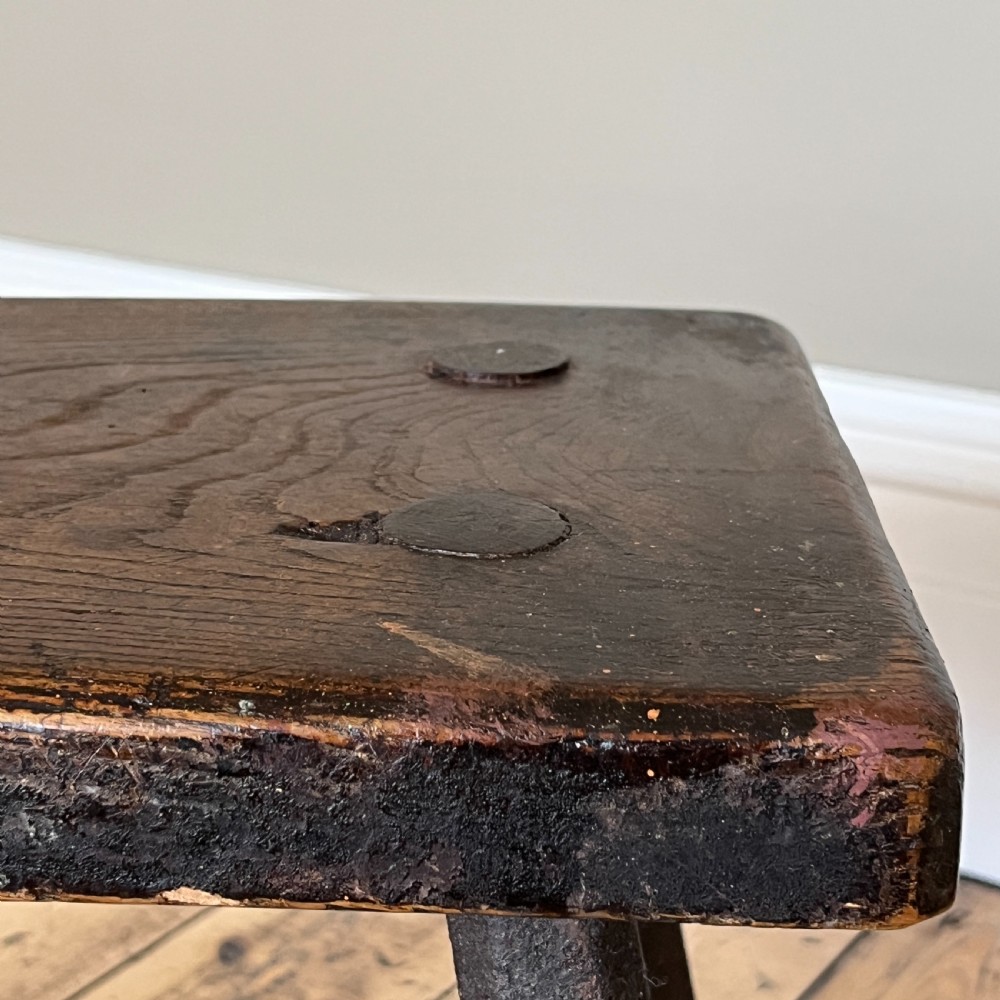 A rustic dark oak four-legged stool featuring a thick rectangular top with four visible circular wedged tenons and splayed faceted legs. View XVII