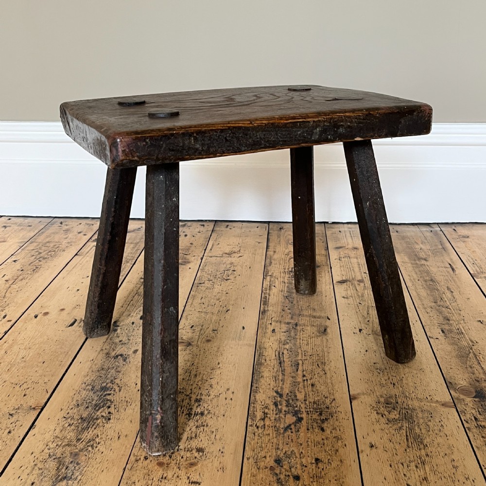 A rustic dark oak four-legged stool featuring a thick rectangular top with four visible circular wedged tenons and splayed faceted legs. View XIX