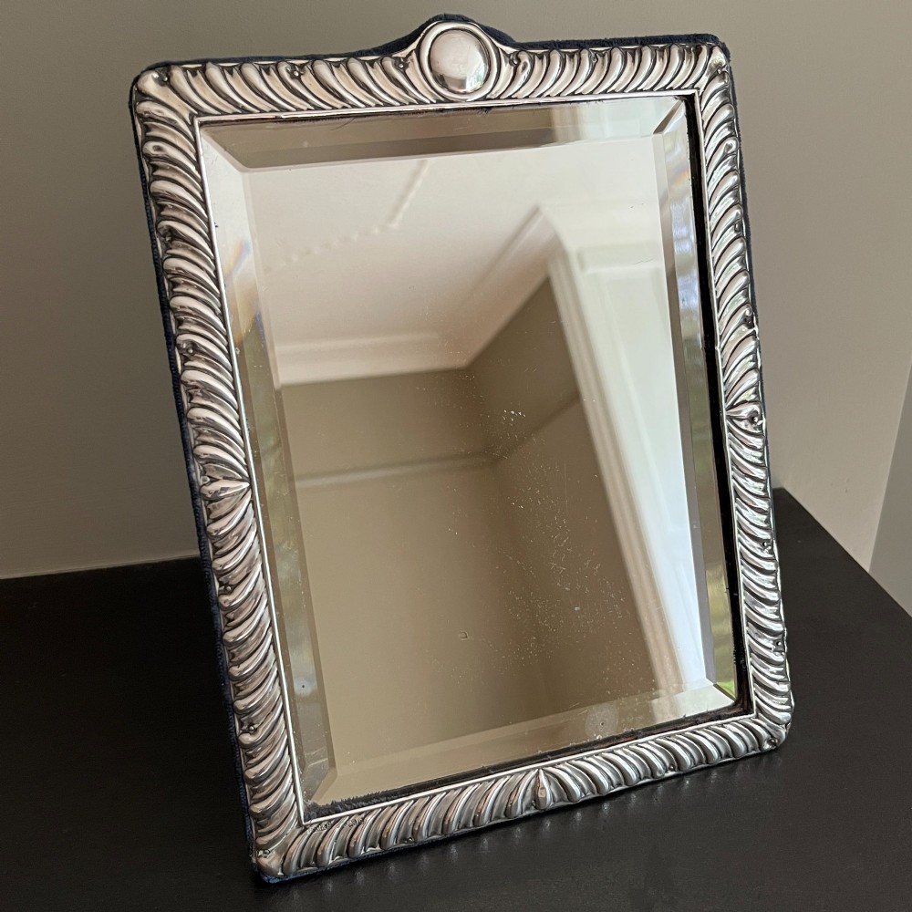 An Edwardian rectangular sterling silver strut mirror featuring a fluted gadrooned frame, a central circular boss crest, and bevelled mirror glass.