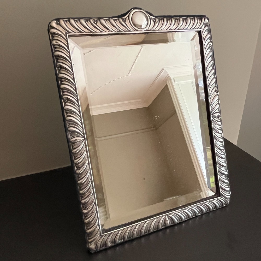 An Edwardian rectangular sterling silver strut mirror featuring a fluted gadrooned frame, a central circular boss crest, and bevelled mirror glass. View II