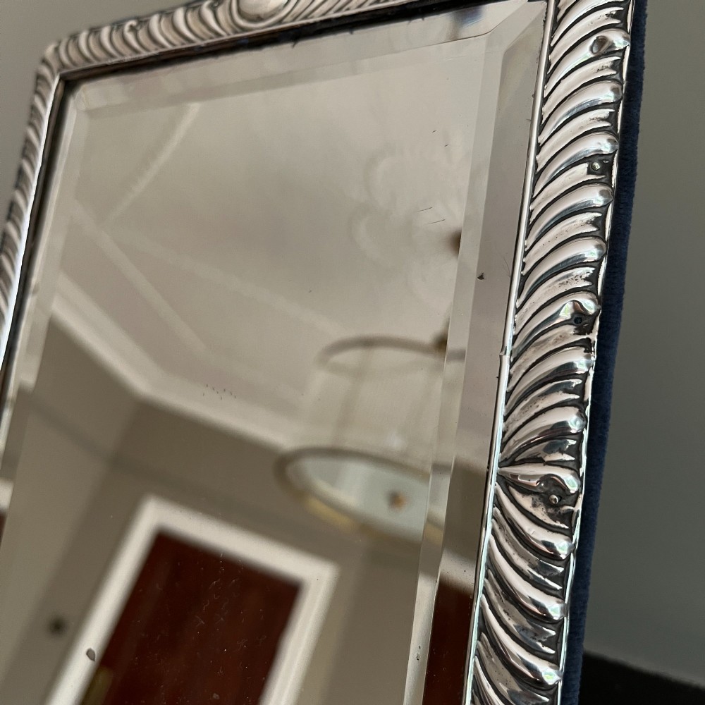 An Edwardian rectangular sterling silver strut mirror featuring a fluted gadrooned frame, a central circular boss crest, and bevelled mirror glass. View V