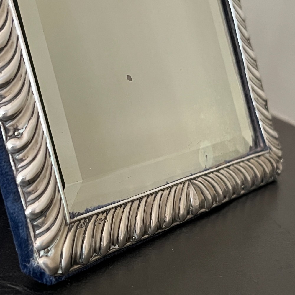 An Edwardian rectangular sterling silver strut mirror featuring a fluted gadrooned frame, a central circular boss crest, and bevelled mirror glass. View VII