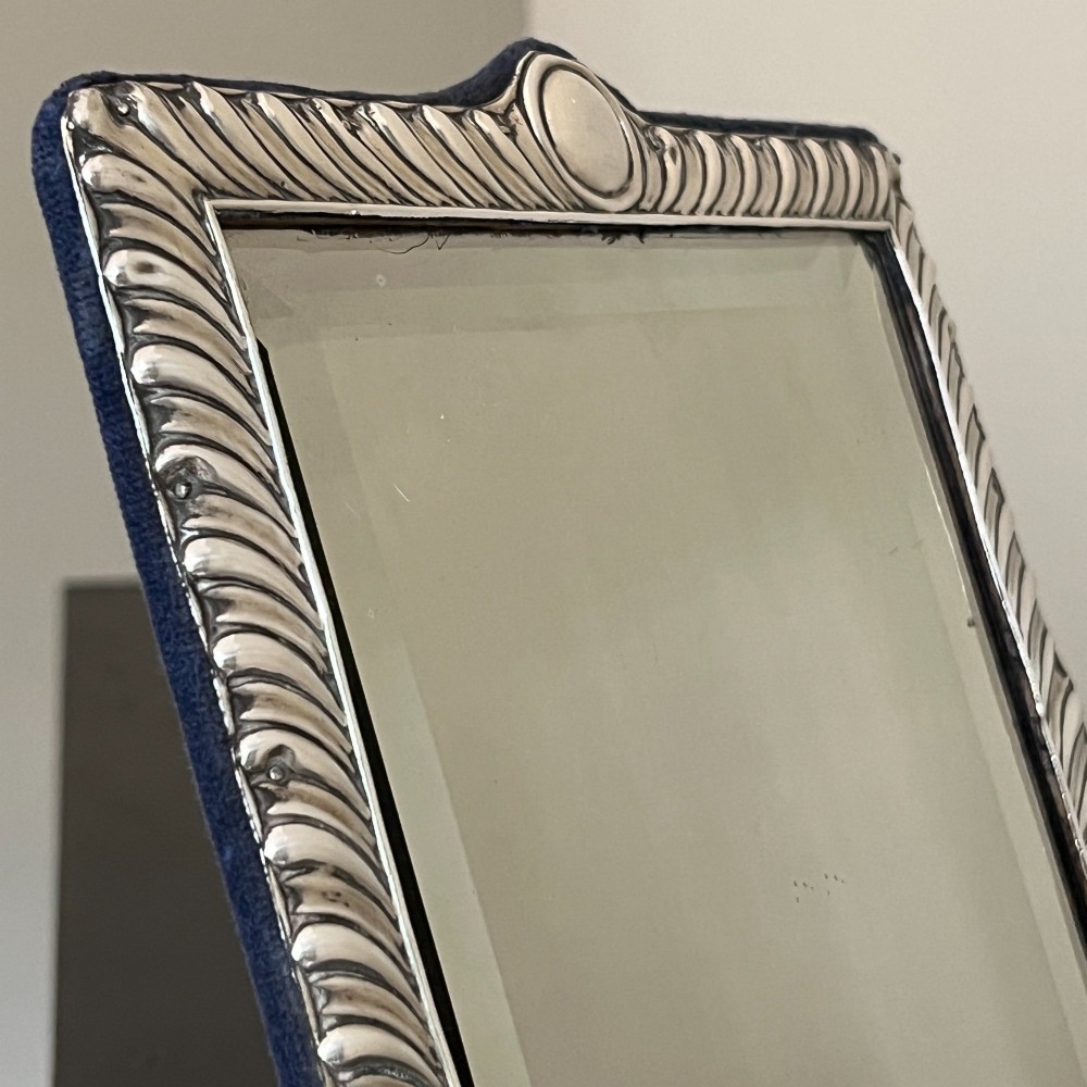 An Edwardian rectangular sterling silver strut mirror featuring a fluted gadrooned frame, a central circular boss crest, and bevelled mirror glass. View IX