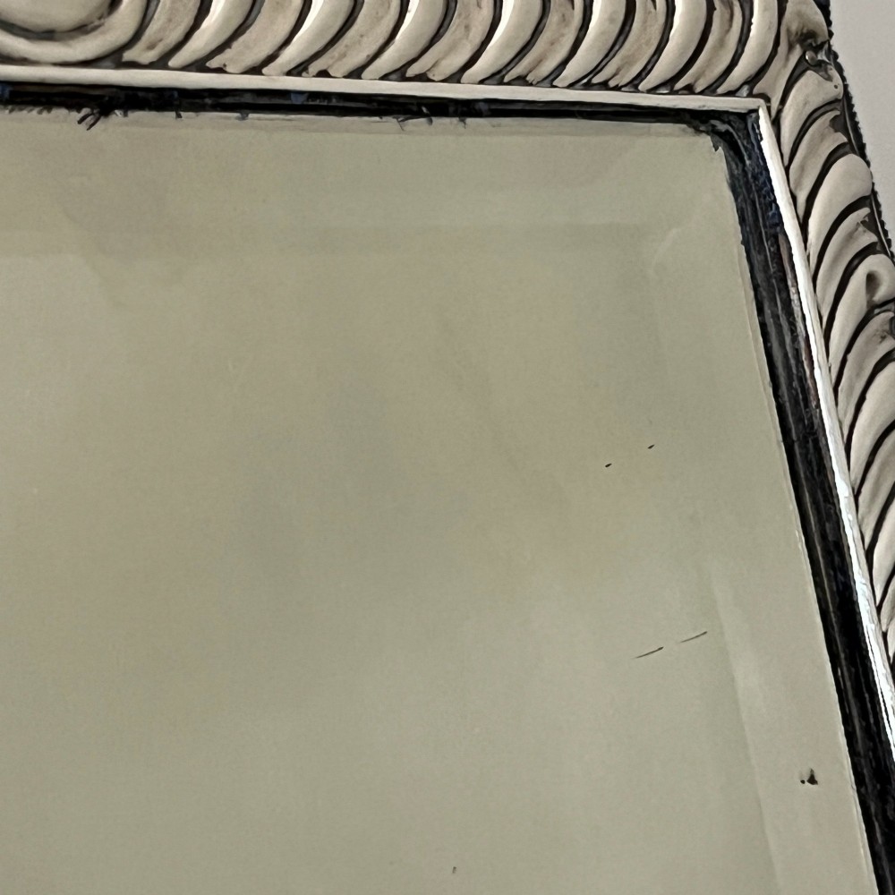 An Edwardian rectangular sterling silver strut mirror featuring a fluted gadrooned frame, a central circular boss crest, and bevelled mirror glass. View XII