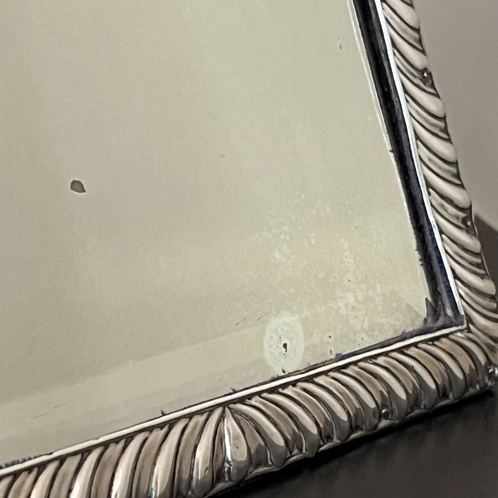 An Edwardian rectangular sterling silver strut mirror featuring a fluted gadrooned frame, a central circular boss crest, and bevelled mirror glass. View XIII