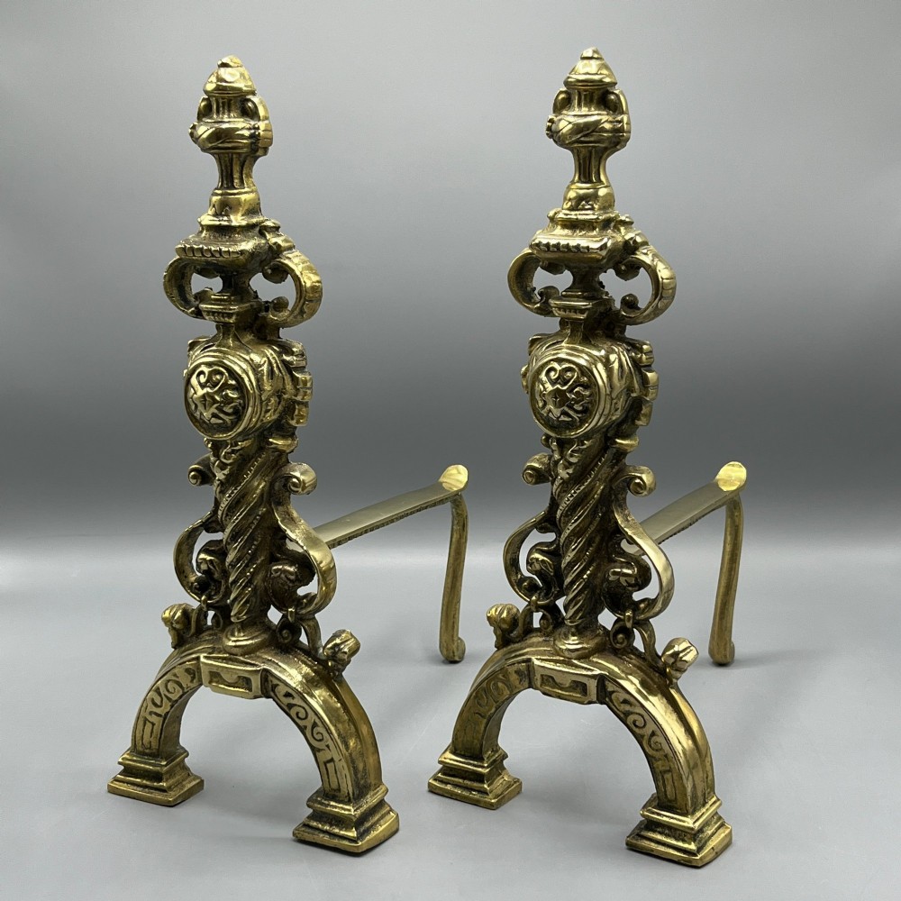 A pair of ornate Victorian solid brass fire dogs featuring finials, twisted columns, medallion details, and arched scrolled feet against a grey backdrop.