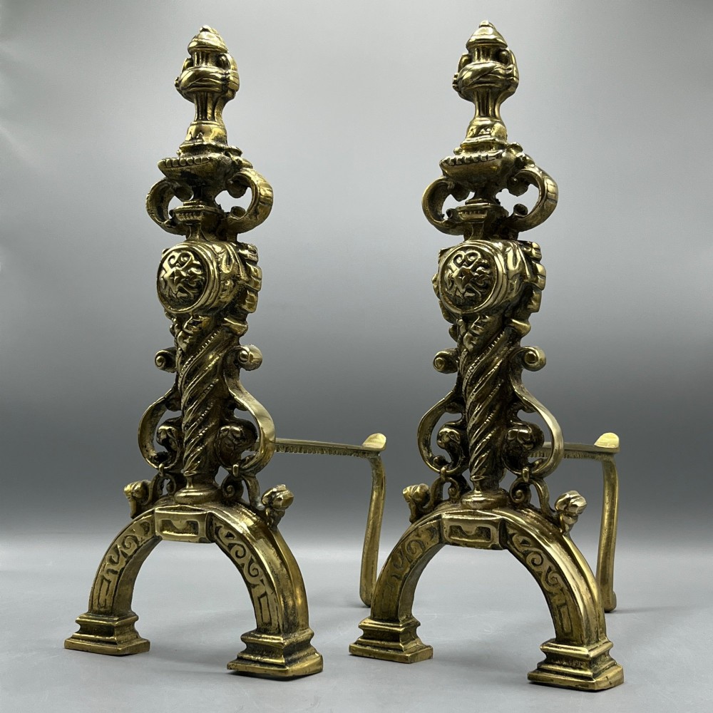 A pair of ornate Victorian solid brass fire dogs featuring finials, twisted columns, medallion details, and arched scrolled feet against a grey backdrop. View II