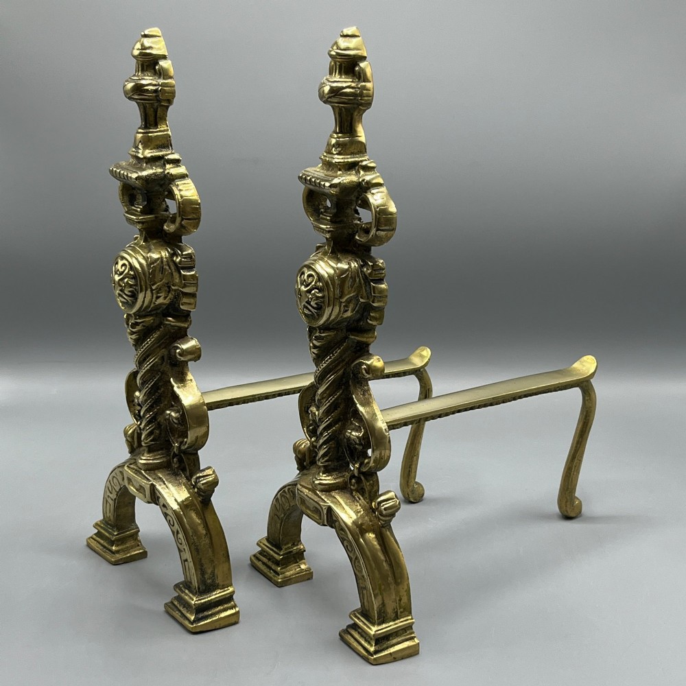 A pair of ornate Victorian solid brass fire dogs featuring finials, twisted columns, medallion details, and arched scrolled feet against a grey backdrop. View III