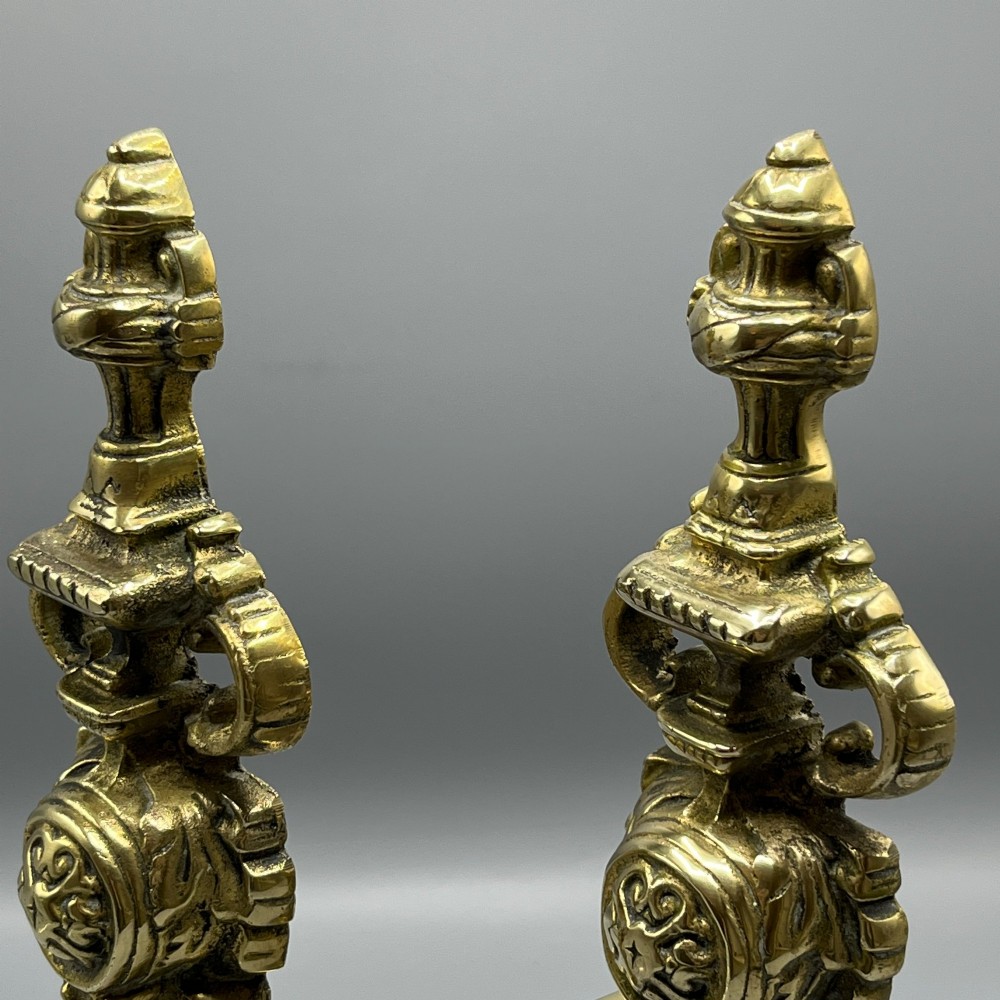 A pair of ornate Victorian solid brass fire dogs featuring finials, twisted columns, medallion details, and arched scrolled feet against a grey backdrop. View IV