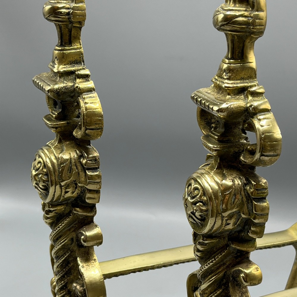 A pair of ornate Victorian solid brass fire dogs featuring finials, twisted columns, medallion details, and arched scrolled feet against a grey backdrop. View V