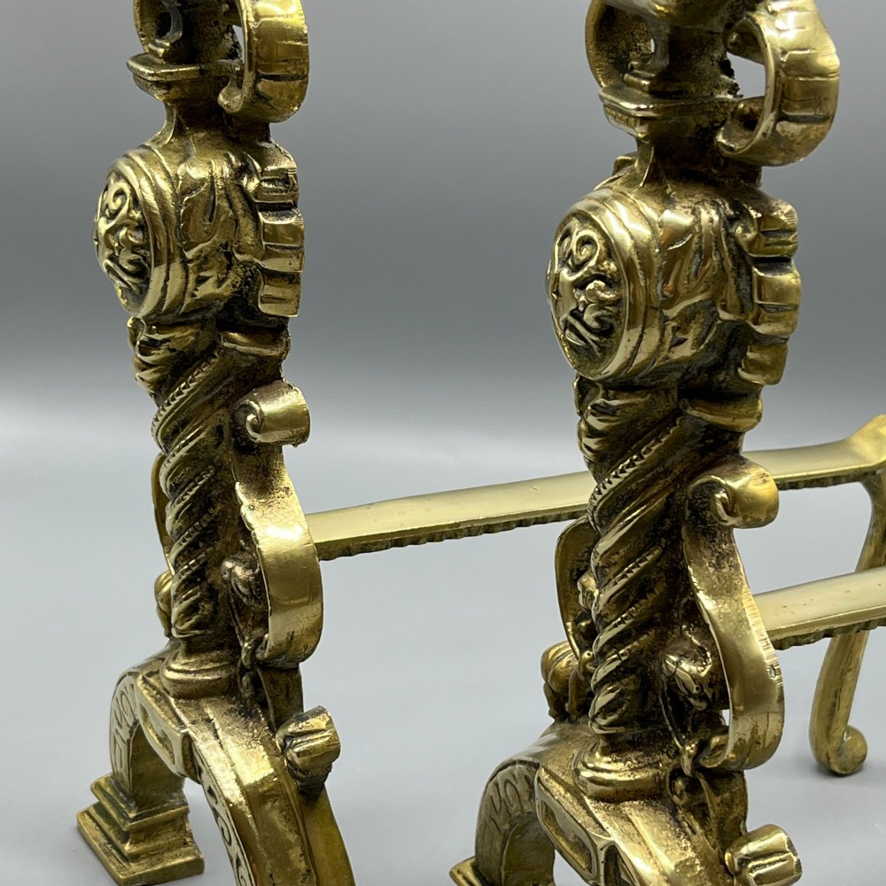 A pair of ornate Victorian solid brass fire dogs featuring finials, twisted columns, medallion details, and arched scrolled feet against a grey backdrop. View VI