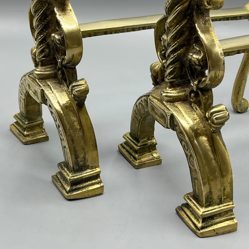 A pair of ornate Victorian solid brass fire dogs featuring finials, twisted columns, medallion details, and arched scrolled feet against a grey backdrop. View VII
