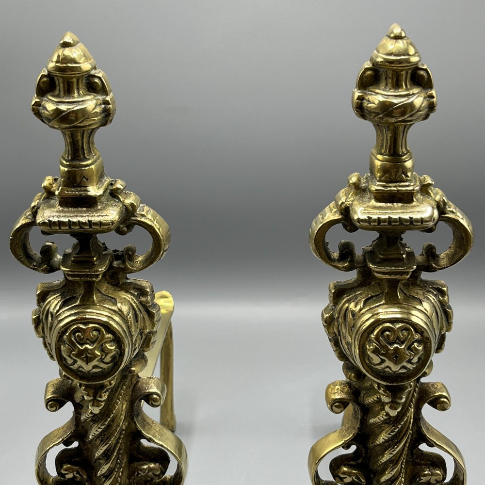 A pair of ornate Victorian solid brass fire dogs featuring finials, twisted columns, medallion details, and arched scrolled feet against a grey backdrop. View VIII