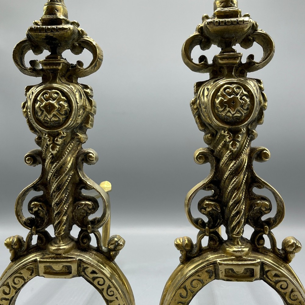 A pair of ornate Victorian solid brass fire dogs featuring finials, twisted columns, medallion details, and arched scrolled feet against a grey backdrop. View IX