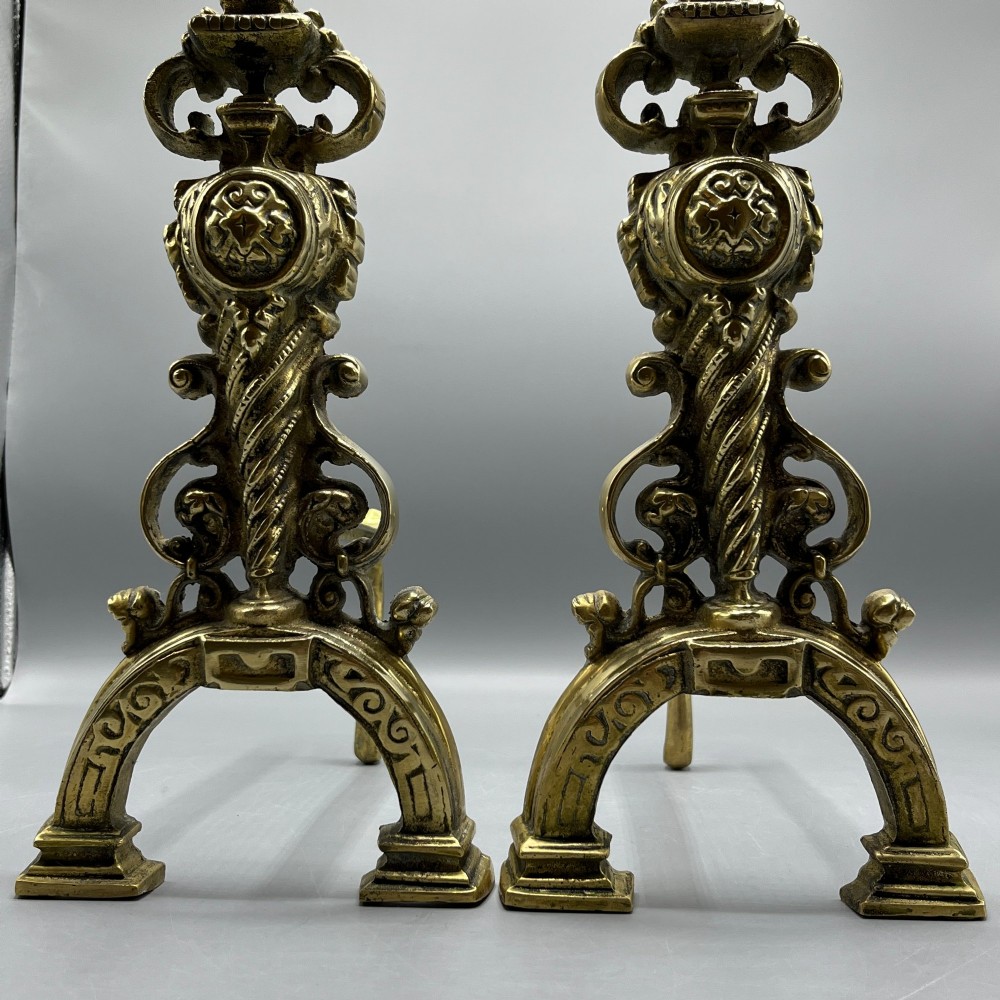 A pair of ornate Victorian solid brass fire dogs featuring finials, twisted columns, medallion details, and arched scrolled feet against a grey backdrop. View X