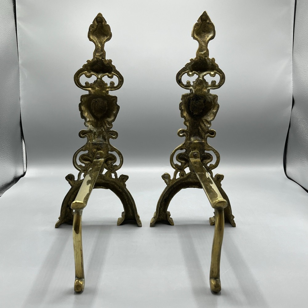 A pair of ornate Victorian solid brass fire dogs featuring finials, twisted columns, medallion details, and arched scrolled feet against a grey backdrop. View XV