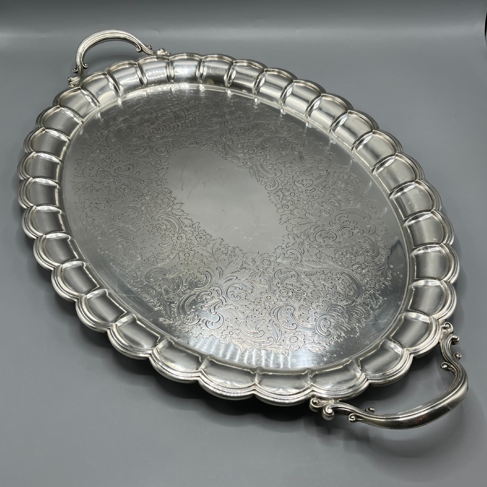 A large oval silver plated serving tray featuring twin scroll handles, a scalloped rim, and chased foliage patterns across the flat interior.