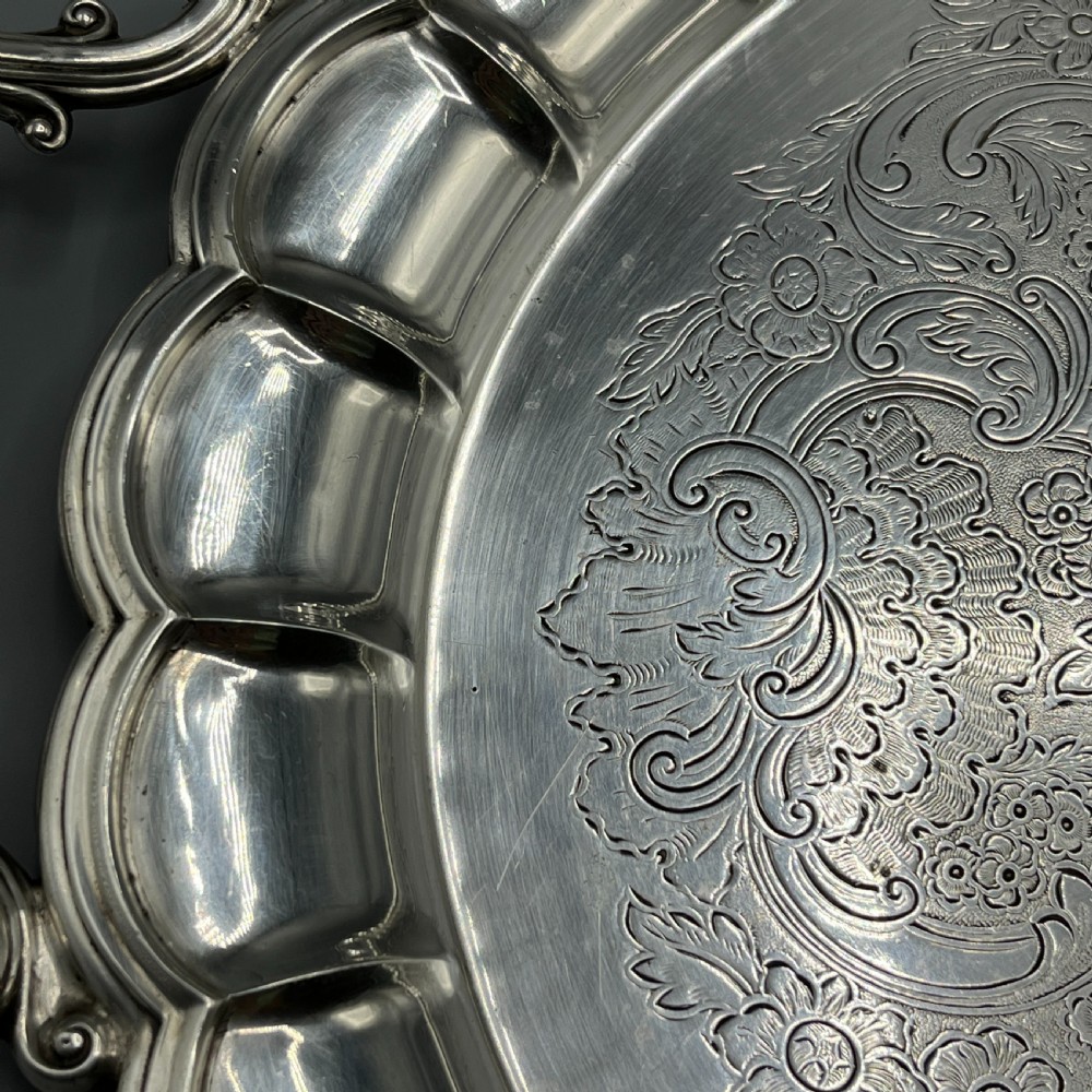 A large oval silver plated serving tray featuring twin scroll handles, a scalloped rim, and chased foliage patterns across the flat interior. View VII