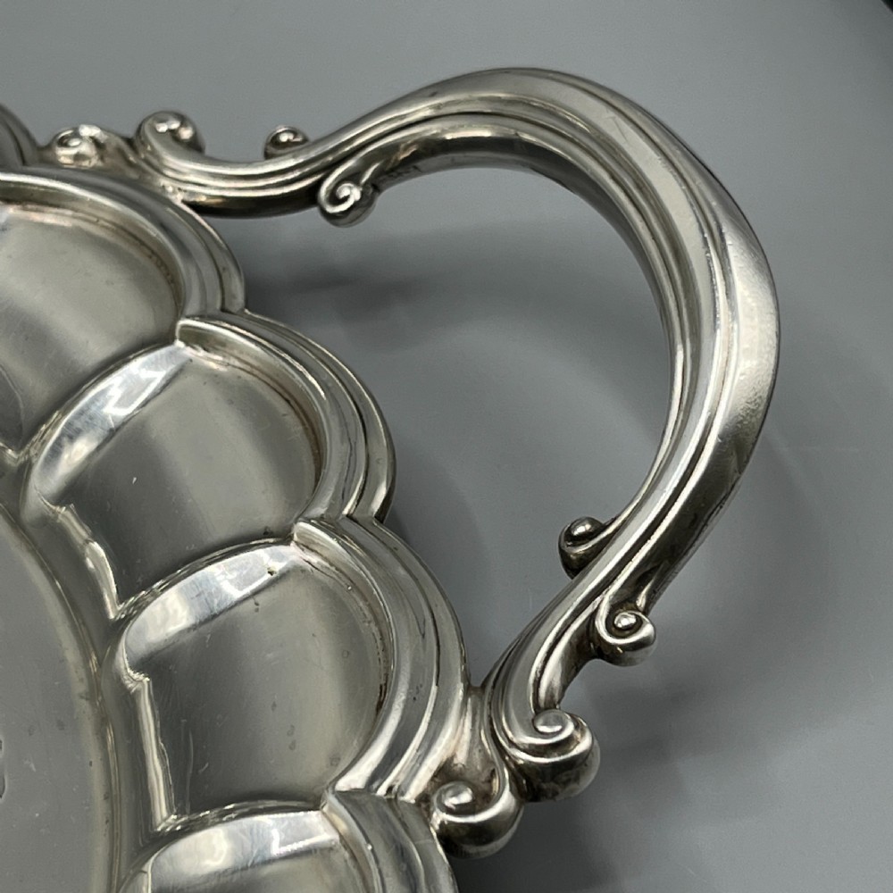 A large oval silver plated serving tray featuring twin scroll handles, a scalloped rim, and chased foliage patterns across the flat interior. View XII