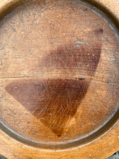 fig 2. A circular turned sycamore wooden platter featuring a raised rim, shallow basin with knife marks, and a dark triangular iron burn mark in the centre.