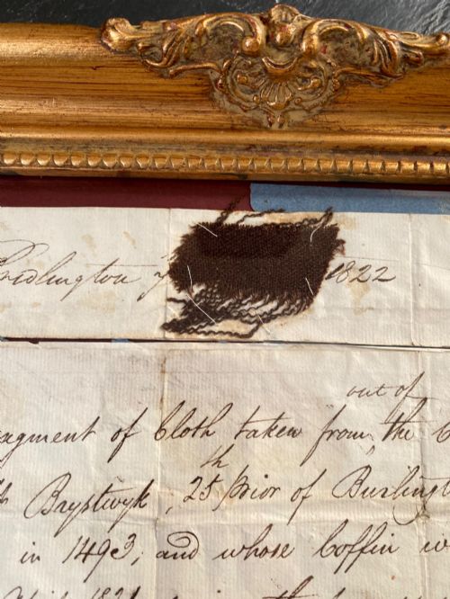 fig 2. A dark 15th-century textile fragment and handwritten 19th-century note mounted inside an ornate carved giltwood frame.