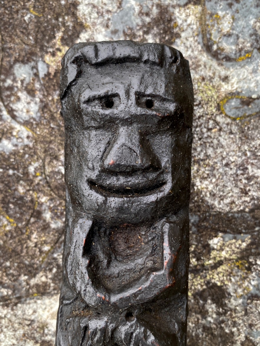 A dark carved oak wooden fragment depicting a stylized human face and decorative geometric relief details. View II