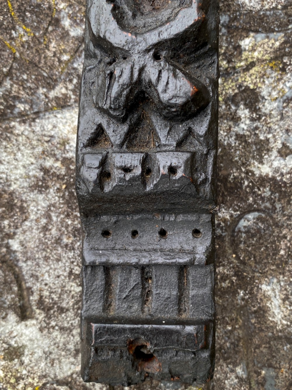 A dark carved oak wooden fragment depicting a stylized human face and decorative geometric relief details. View VII