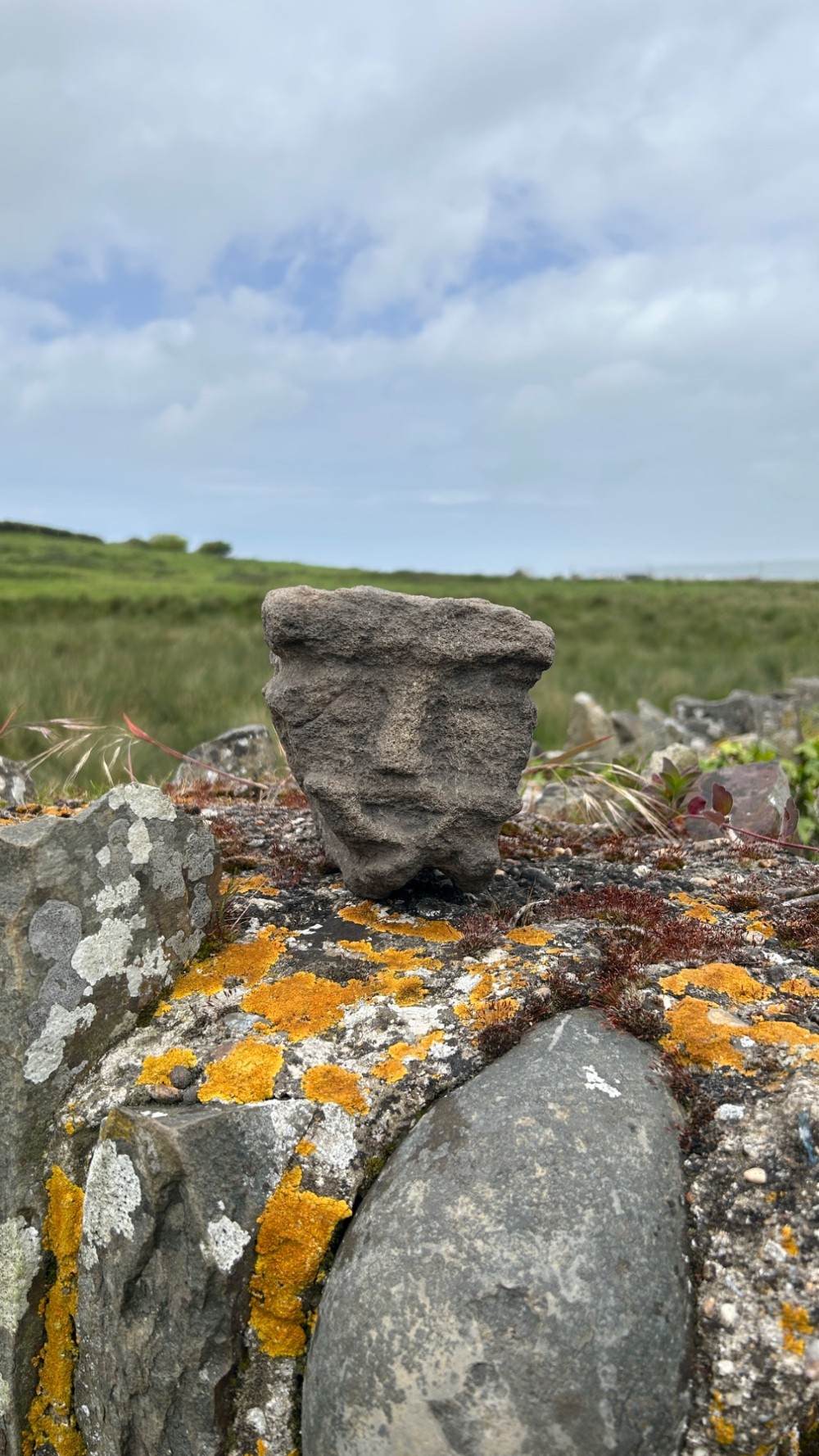 A hand-carved medieval grey stone corbel featuring a stylised human face with a gripping mouth detail. View V