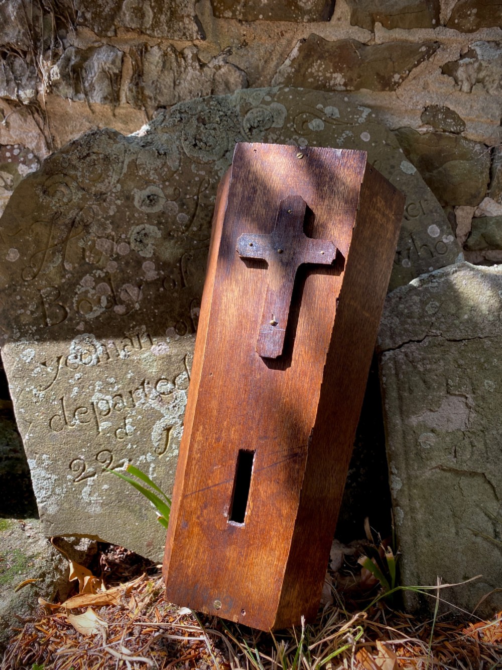 A wooden coffin-shaped collection box featuring a raised wooden cross motif and a narrow coin slot on the front lid.