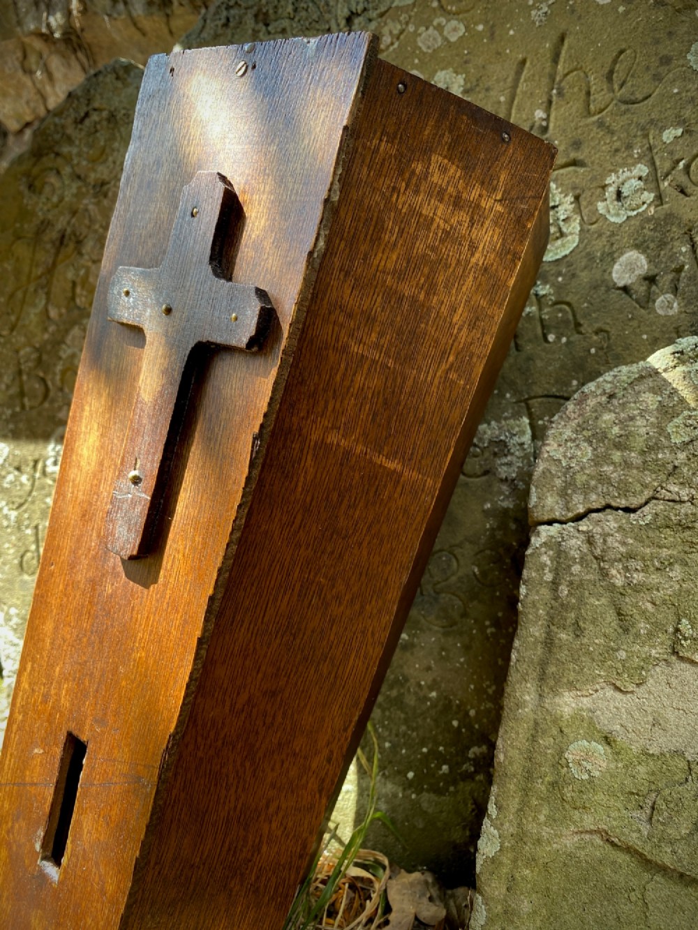 A wooden coffin-shaped collection box featuring a raised wooden cross motif and a narrow coin slot on the front lid. View VI
