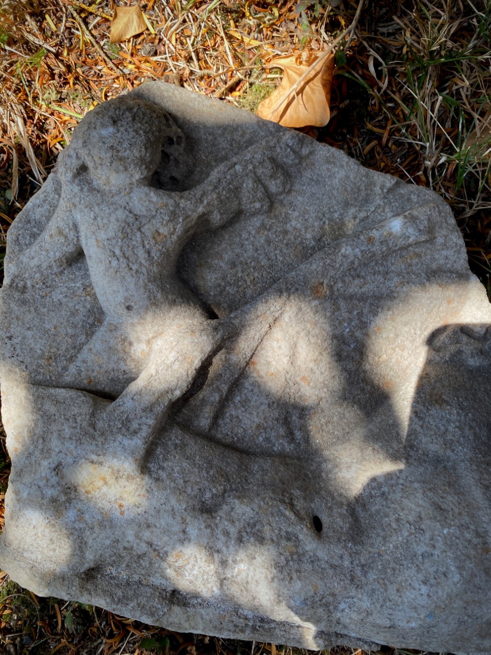 A carved weathered stone fragment depicting a high-relief figure of a winged cherub above a reclining form. View X