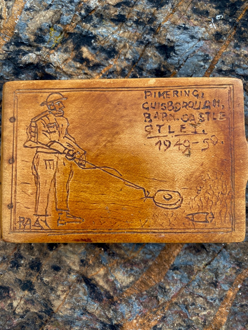 A rectangular carved wooden box lid depicting a uniformed military figure with a detector alongside hand-incised lettering detailing UK locations and dates.