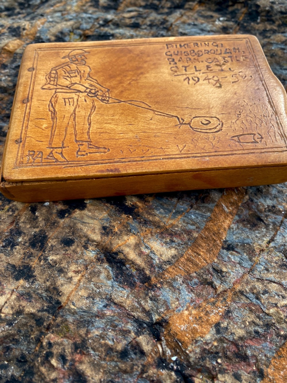 A rectangular carved wooden box lid depicting a uniformed military figure with a detector alongside hand-incised lettering detailing UK locations and dates. View IV