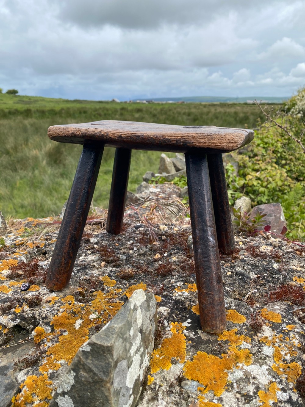 A low rectangular wooden hearth stool featuring dark aged oak timber, four splayed legs, and exposed wedged tenon joints on the top surface. View III