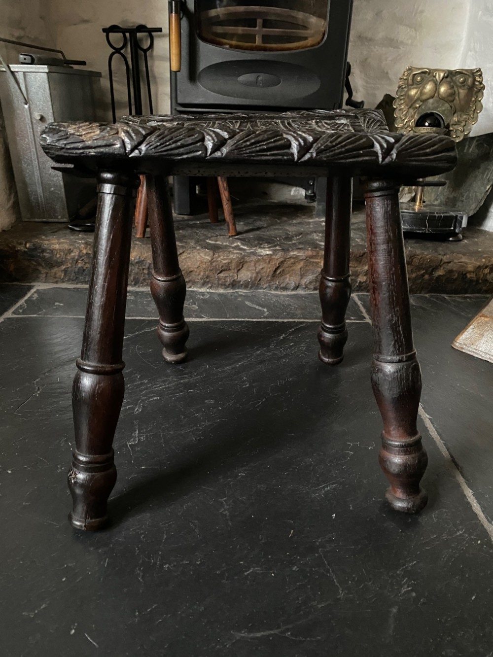 A dark brown carved oak stool featuring hand-carved shell motif decoration on the top seat and four turned wooden legs.