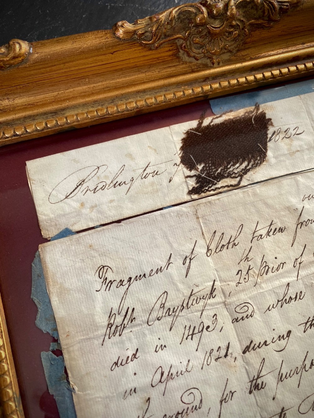 A dark 15th-century textile fragment and handwritten 19th-century note mounted inside an ornate carved giltwood frame. View II