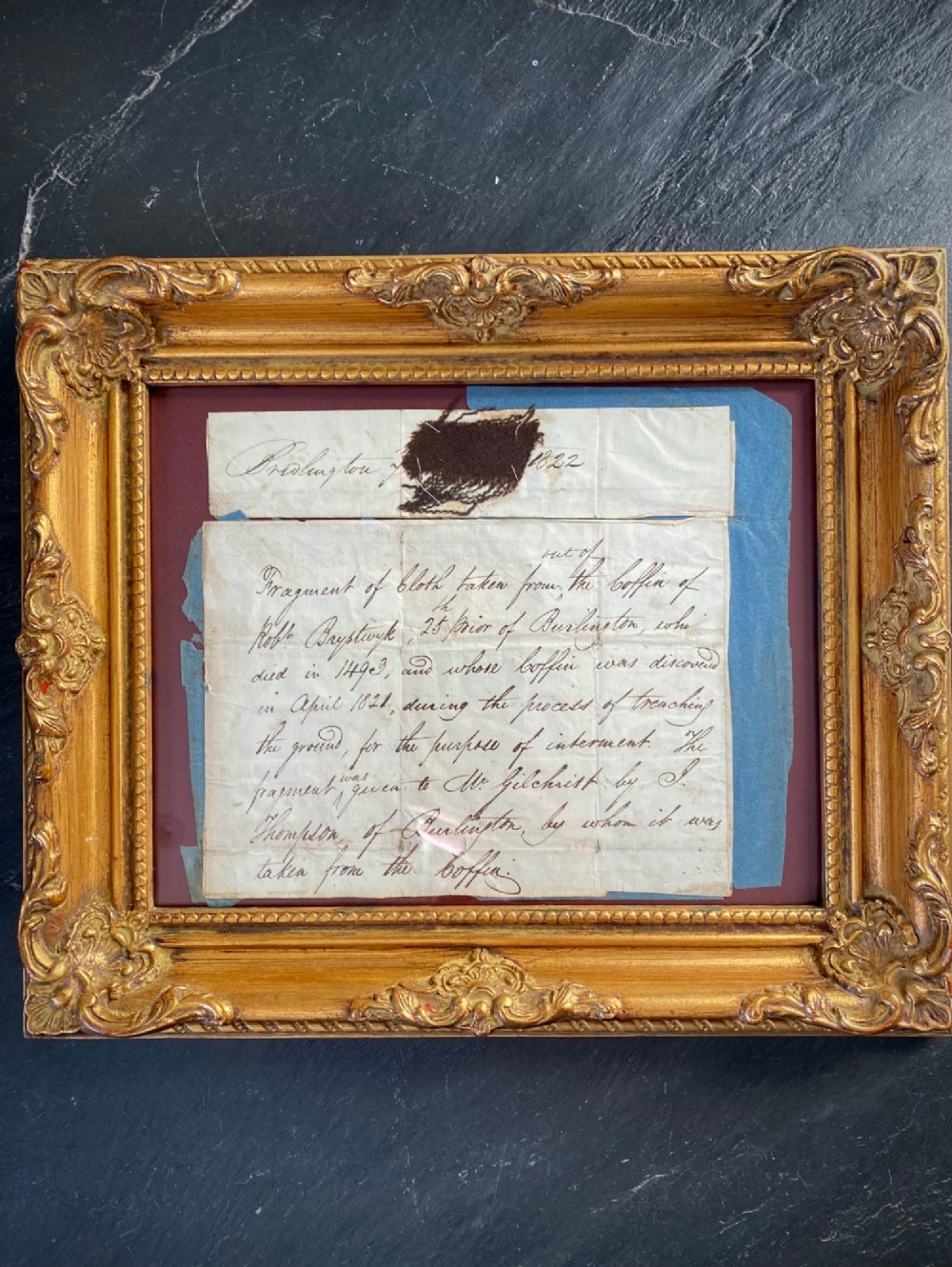A dark 15th-century textile fragment and handwritten 19th-century note mounted inside an ornate carved giltwood frame.