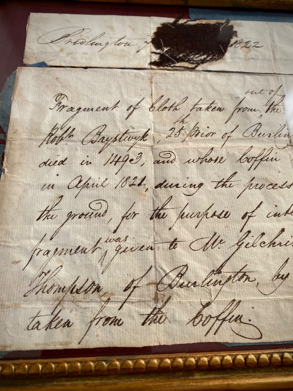 A dark 15th-century textile fragment and handwritten 19th-century note mounted inside an ornate carved giltwood frame. View IV