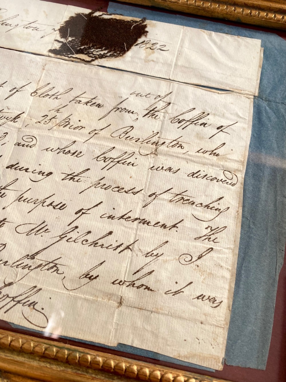 A dark 15th-century textile fragment and handwritten 19th-century note mounted inside an ornate carved giltwood frame. View V