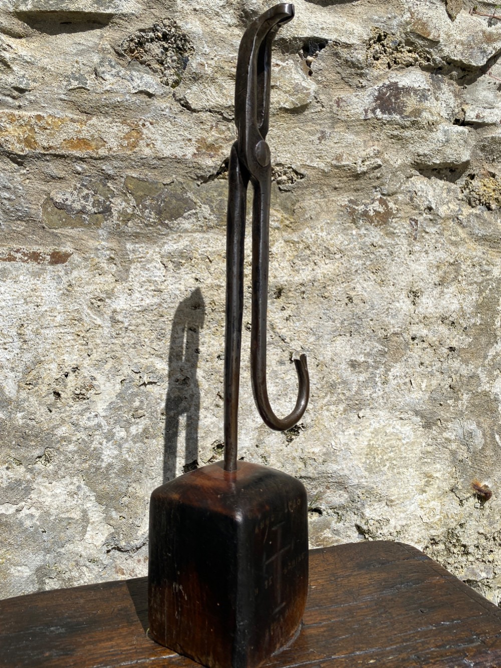A forged steel tool with an integral lower hook mounted vertically into a square dark wooden block engraved with a cross and text.