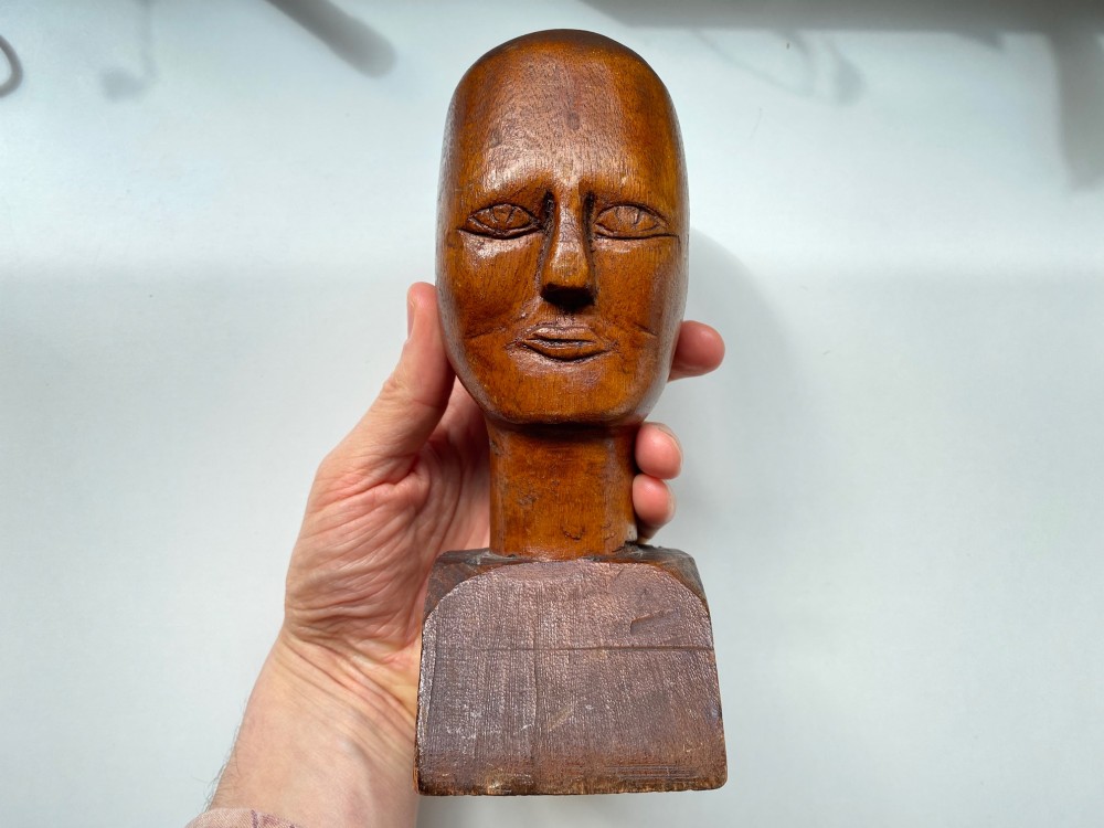 A small hand-carved wooden head sculpture on a square base with stylized eyes and mouth in a warm brown finish.
