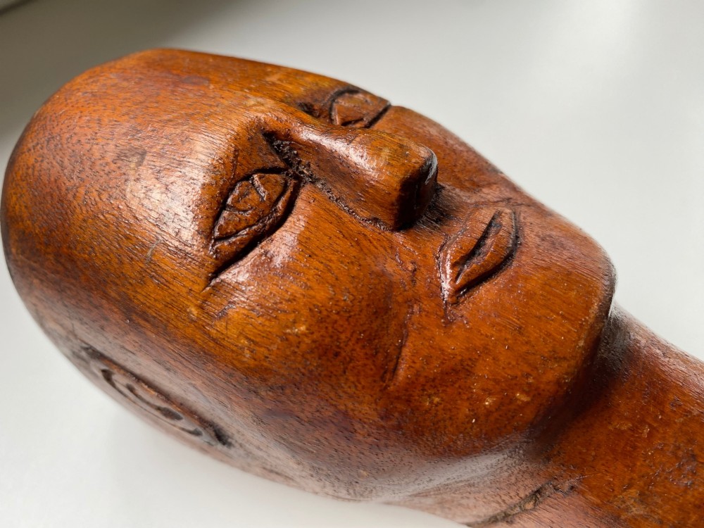 A small hand-carved wooden head sculpture on a square base with stylized eyes and mouth in a warm brown finish. View II