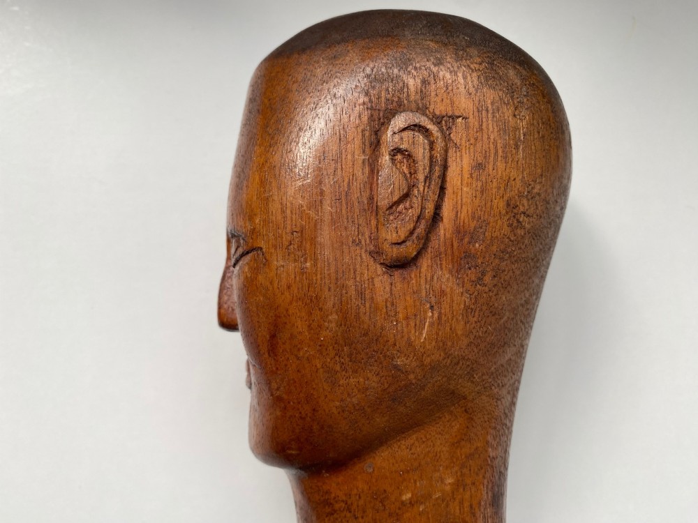 A small hand-carved wooden head sculpture on a square base with stylized eyes and mouth in a warm brown finish. View III